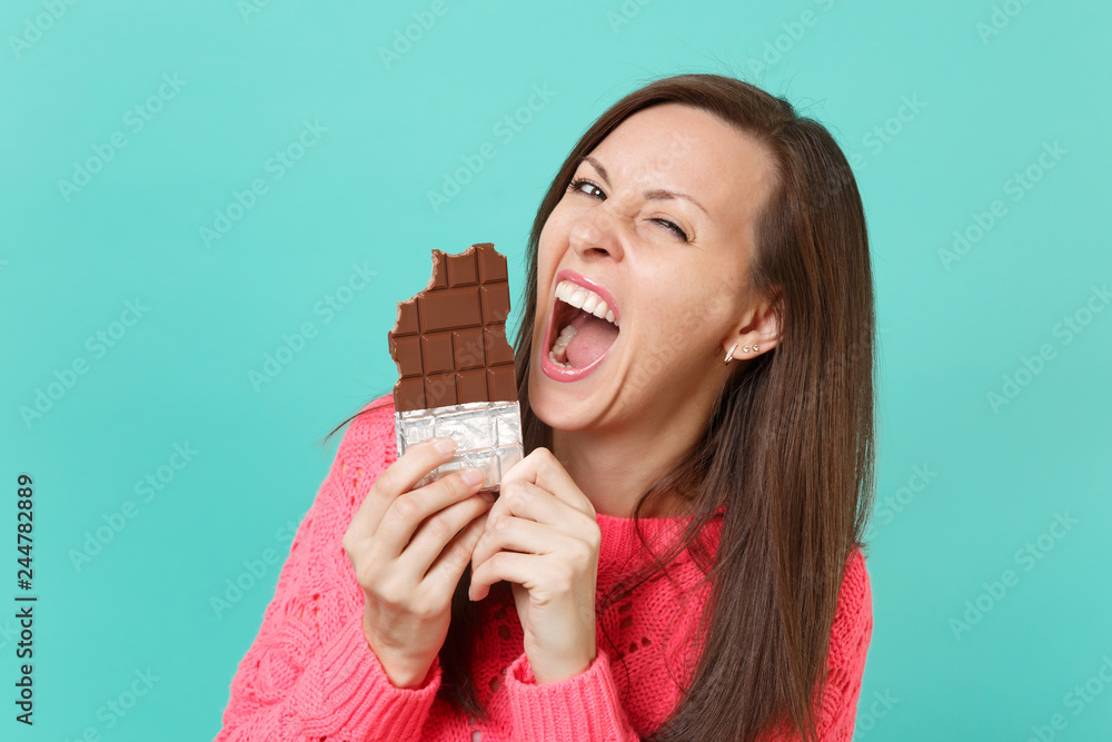 Crazy cheerful young woman in knitted pink sweater holding in hand and biting chocolate bar isolated on blue turquoise wall background, studio portrait. People lifestyle concept. Mock up copy space. Crazy cheerful young woman in knitted pink sweater holding in hand and biting chocolate bar isolated on blue turquoise wall background, studio portrait. People lifestyle concept. Mock up copy space.