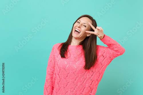 Laughing attractive young woman in knitted pink sweater showing victory sign isolated on blue turquoise wall background studio portrait. People sincere emotions  lifestyle concept. Mock up copy space.