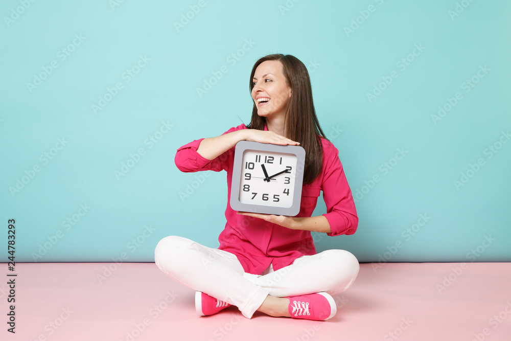 Full length portrait young woman in rose shirt blouse white pants sitting on floor hold clock isolated on bright pink blue pastel wall background studio. Fashion lifestyle concept. Mock up copy space. Full length portrait young woman in rose shirt blouse white pants sitting on floor hold clock isolated on bright pink blue pastel wall background studio. Fashion lifestyle concept. Mock up copy space.