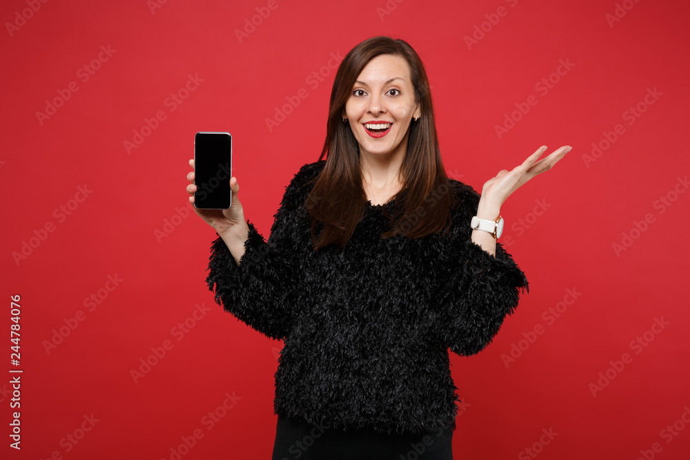 Naklejka premium Amazed young woman in black fur sweater spreading hand, holding mobile phone with blank empty screen isolated on bright red background. People sincere emotions, lifestyle concept. Mock up copy space.