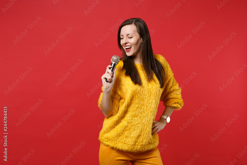 Portrait of stunning pretty young woman in yellow fur sweater sing song in microphone isolated on bright red wall background in studio. People sincere emotions, lifestyle concept. Mock up copy space. Portrait of stunning pretty young woman in yellow fur sweater sing song in microphone isolated on bright red wall background in studio. People sincere emotions, lifestyle concept. Mock up copy space.