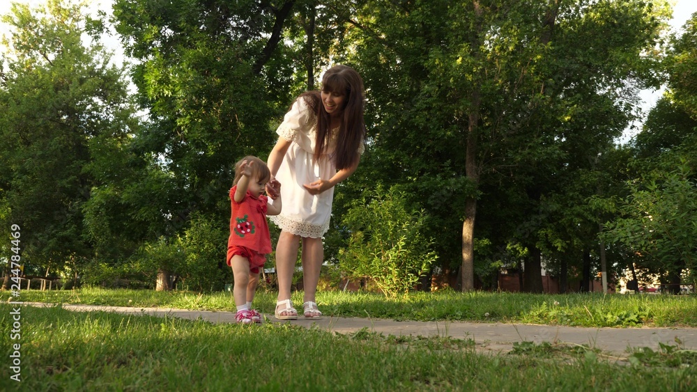 Mom and little daughter are walking along path in summer park. baby's first steps with mom