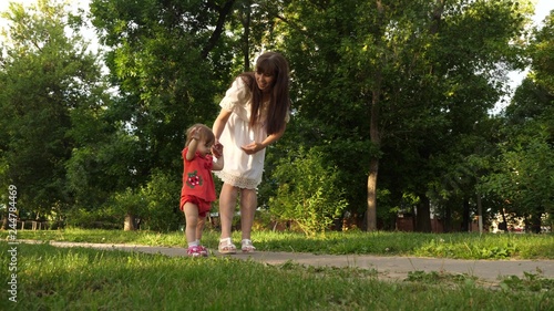 Mom and little daughter are walking along path in summer park. baby's first steps with mom