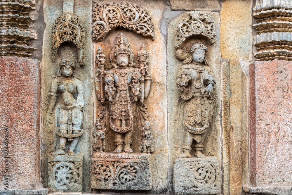 Belur, Karnataka, India - November 2, 2013: Brown wall stone panel sculpture of Lord Vishnu surrouned by his two consorts Devi Lakshmi and Devi Bhu with Shilabalika women. On side of temple building.