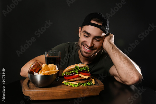 Young man and tasty burger served on wooden board with French fries against black background