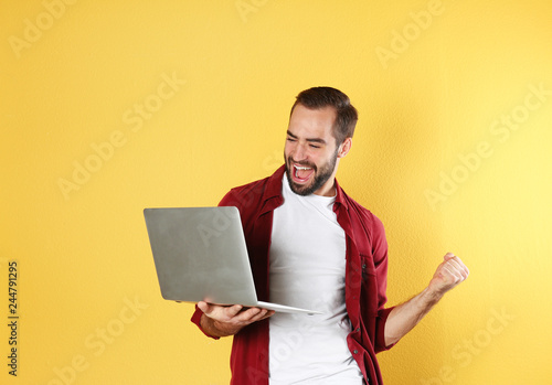 Emotional young man with laptop celebrating victory on color background