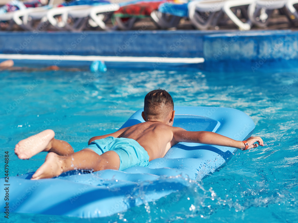 Smiling European boy is swimming on the inflatable blue floater in the hotel’s pool. Back view.