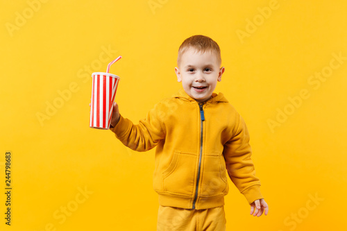 Little fun cheerful kid boy 3-4 years old wearing yellow clothes hold cup of soda isolated on orange wall background, children studio portrait. People, childhood lifestyle concept. Mock up copy space.