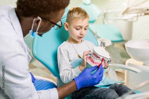 African-American ethnic black male dentist in blue latex gloves checking condition of teeth of little smiling boy. Little boy in blue dental chair in modern dentistry clinic