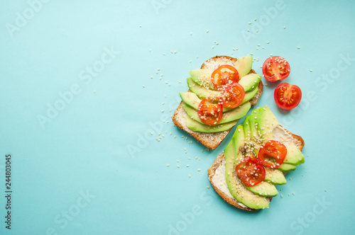 Flat lay of delicious toasts with sliced avocado, tomatoes and sesamum seeds on blue background with copyspace.