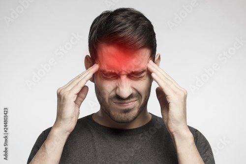 Portrait of young man isolated on gray background, suffering from severe headache, pressing fingers to temples with closed eyes photo