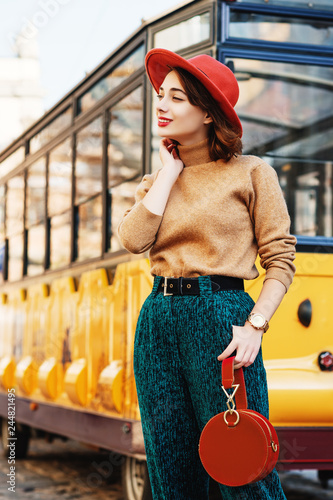 Outdoor portrait of young beautiful fashionable woman wearing orange hat, beige turtleneck, belt, green corduroy trousers, wrist watch, holding round suede handbag, posing in street of european city