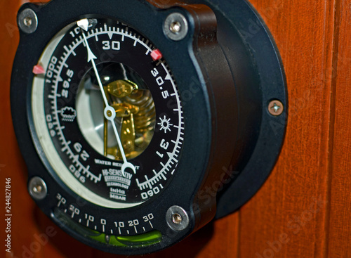 Close up of black ship's barometer with white numbers and pointer, gold mechanism, green ball level, big silver nuts, and white and red markers hanging on crackled teak orange wainscoting background. photo
