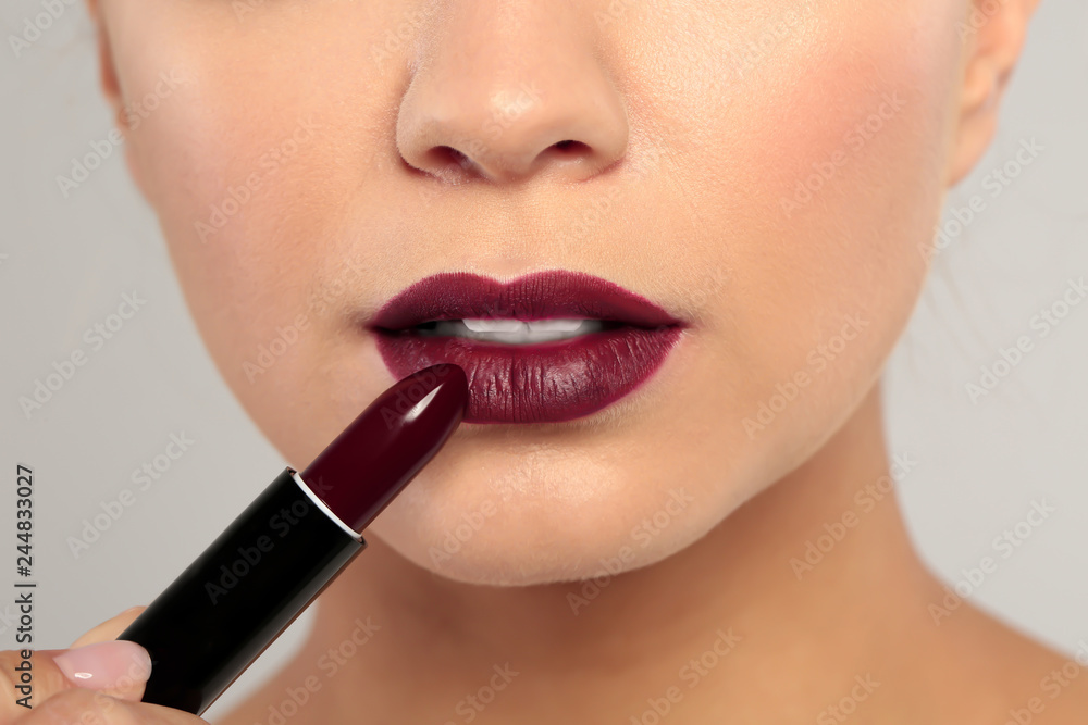 Beautiful woman applying dark lipstick on gray background, closeup