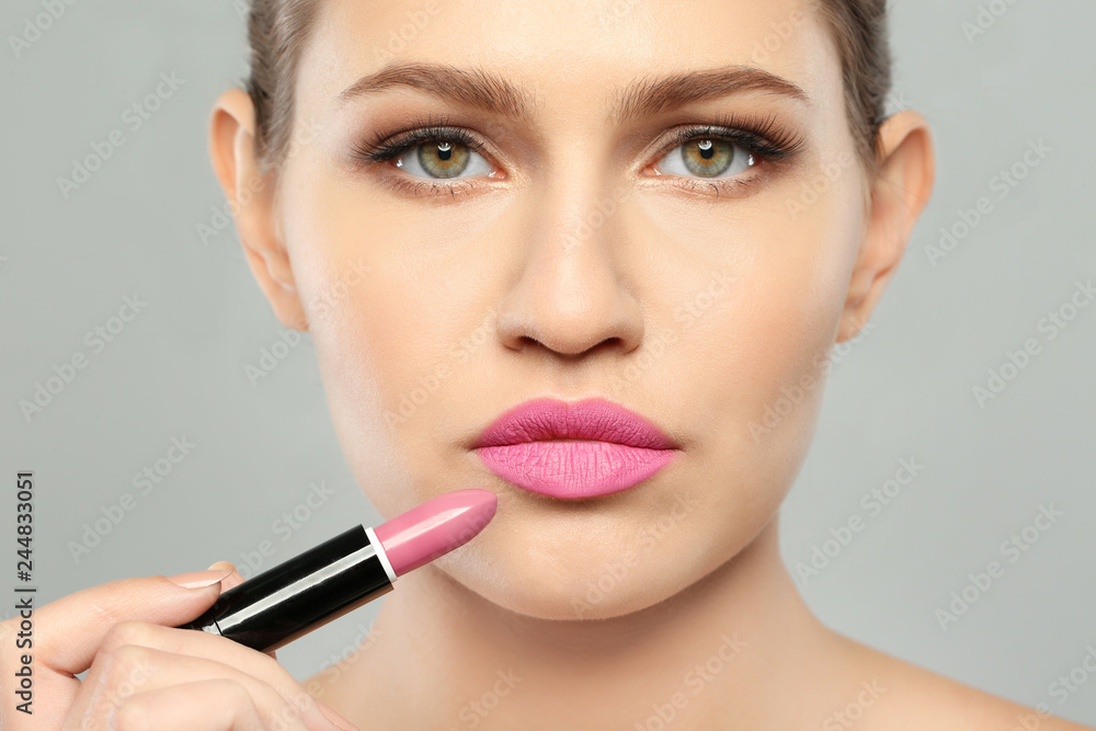 Young woman applying beautiful lipstick on gray background