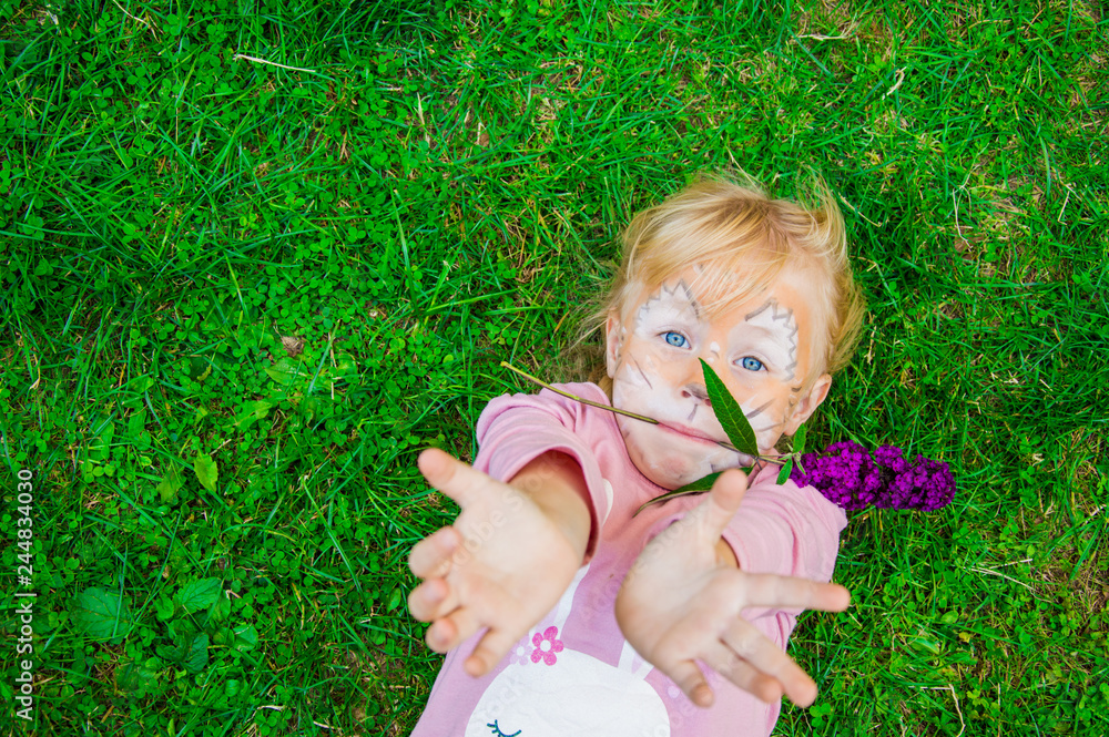 beautiful little girl blonde on a children's holiday with a pattern of a lion cub on her face, emotionally posing while lying on bright green grass holding a flower in her teeth beautiful little girl blonde on a children's holiday with a pattern of a lion cub on her face, emotionally posing while lying on bright green grass holding a flower in her teeth