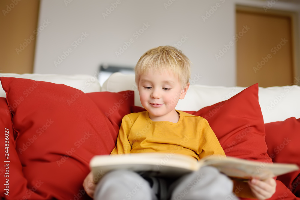 Fototapeta premium Little boy is sitting at home on the couch and reading a book. Learning to read.