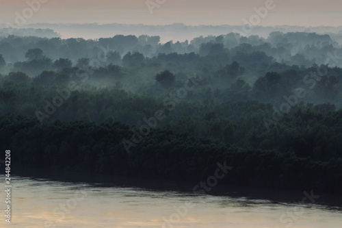 Mystical view on riverbank of large island with forest under haze at early morning. Mist among layers from tree silhouettes under warm predawn sky. Morning atmospheric landscape of majestic nature.