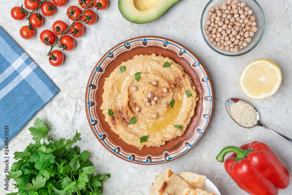 Hummus in to a brown clay plate with a blue pattern. On the white table are vegetables, greens, triangular pieces of pita. Top view. Flat lay. Hummus in to a brown clay plate with a blue pattern. On the white table are vegetables, greens, triangular pieces of pita. Top view. Flat lay.