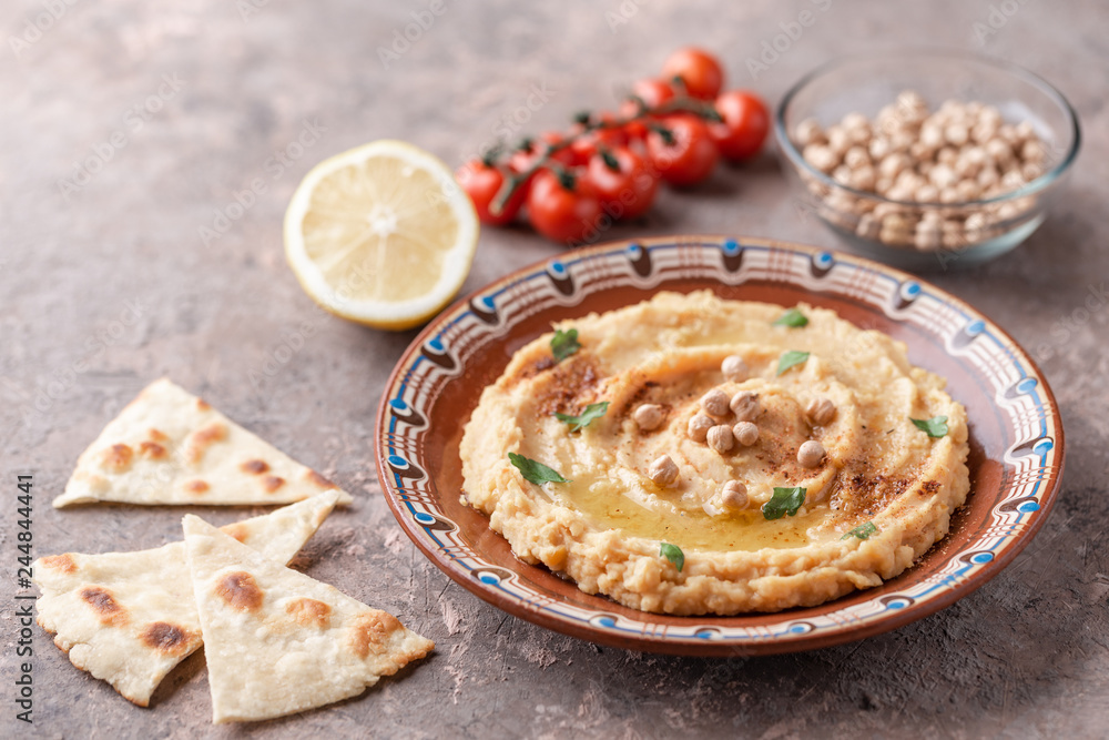 Hummus in a brown clay plate with a blue pattern. On the brown table are vegetables, greens, triangular pieces of pita. Hummus in a brown clay plate with a blue pattern. On the brown table are vegetables, greens, triangular pieces of pita.