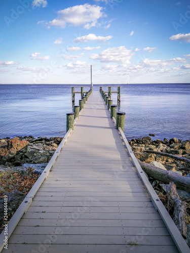 Colorful, Serene Photo of a Dock Extending Out To the Ocean - with a Patch of Rocks and Logs, and Calm Waters on a Bright, Mildly Cloudy Day in the Mid Atlantic United States