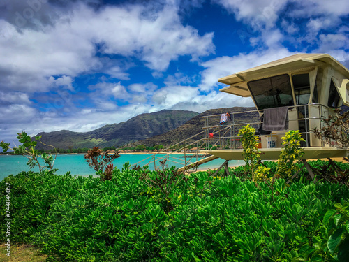 Lifeguard Station in Green Brush - Looking Out Over Aqua Colored Water on the Beach with a Cloudy Blue Sky and Mountains in the Background, in Hawaii on a Bright Spring Day. © Jon