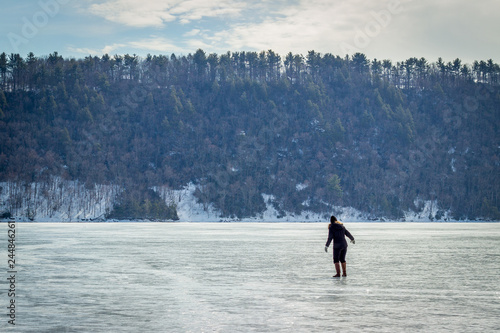Woman in Black Walking on Top of a Frozen Lake - with a Hill and Trees in the Background on a Cold, Winter Day