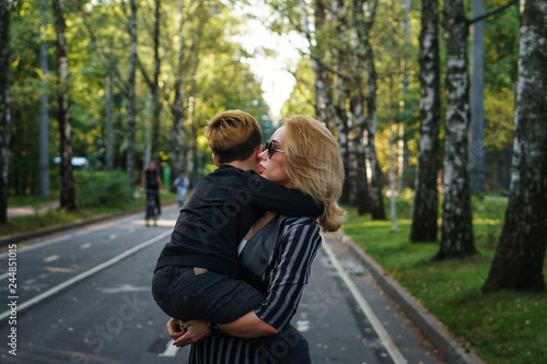 Young attractive mother holding her son in her arms. Blond woman in sunglasses. They walk outdoors in park.