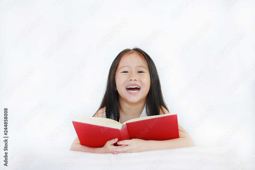 Happy little Asian kid girl reading hardcover book lying with pillow on bed against white background. Happy little Asian kid girl reading hardcover book lying with pillow on bed against white background.