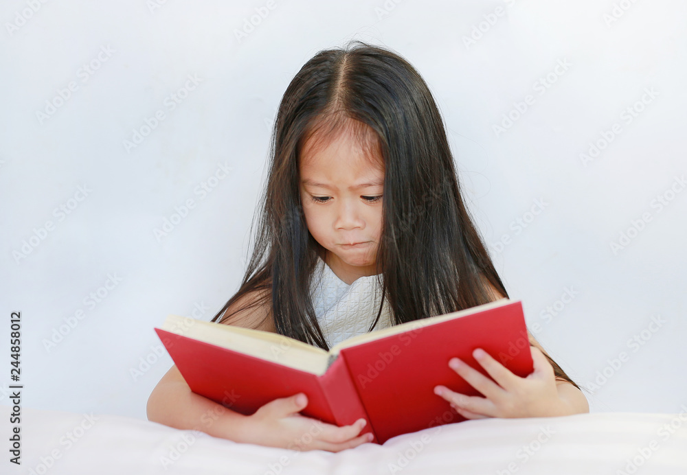 Beautiful little Asian child girl reading hardcover book lying with pillow on white background. Beautiful little Asian child girl reading hardcover book lying with pillow on white background.