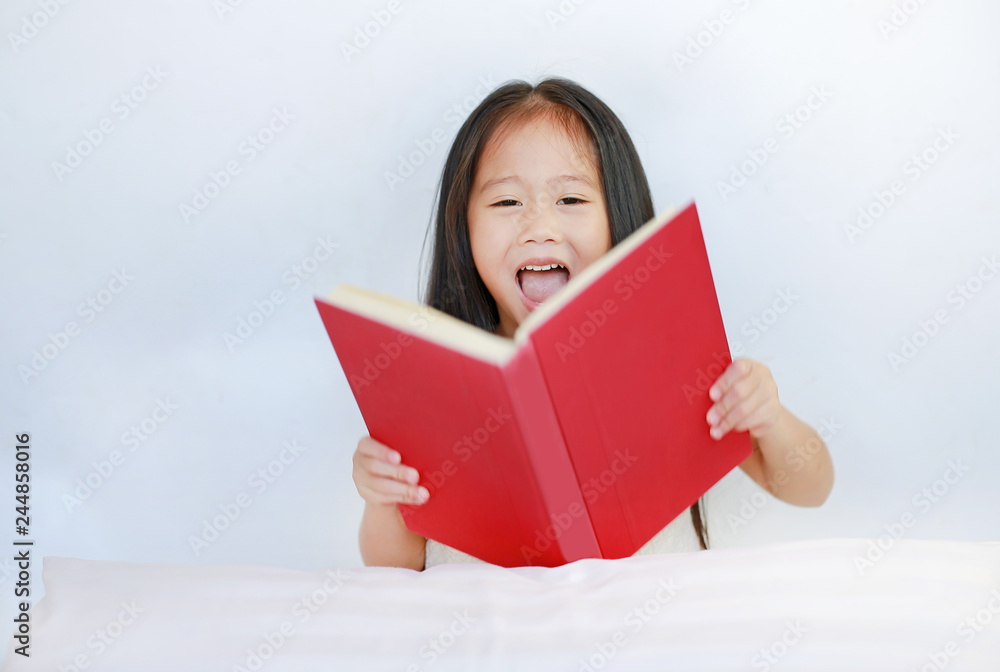 Smiling little Asian kid girl reading hardcover book lying with pillow on bed against white background. Smiling little Asian kid girl reading hardcover book lying with pillow on bed against white background.