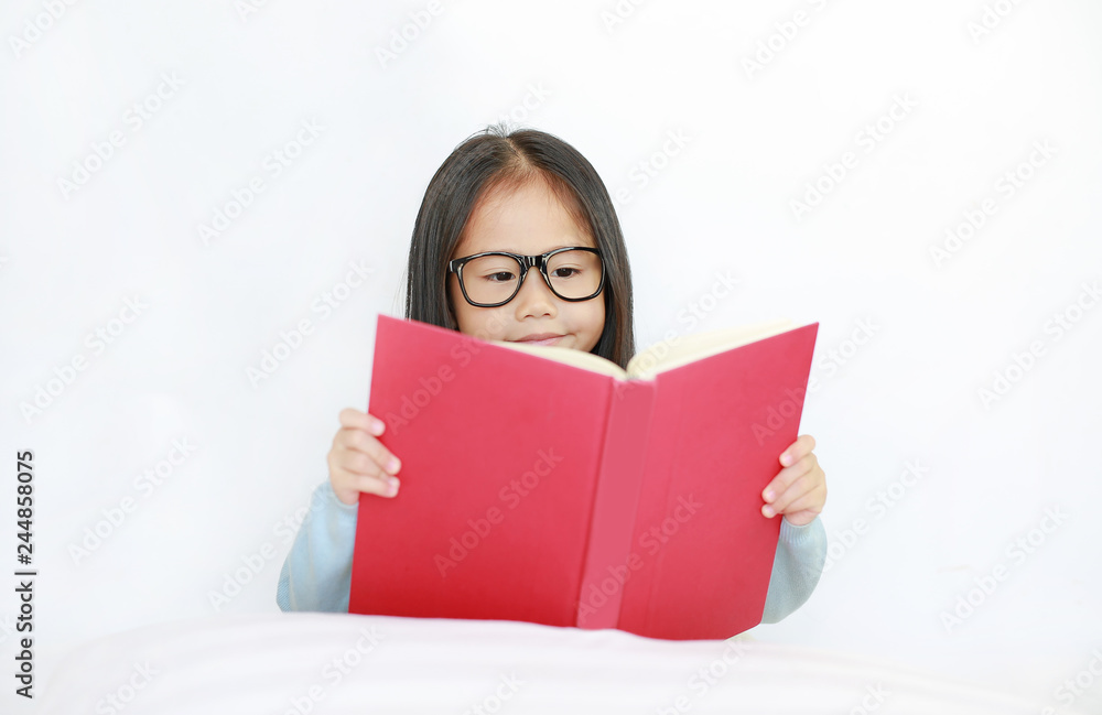 Beautiful happy little Asian kid girl reading hardcover book lying on bed against white background.