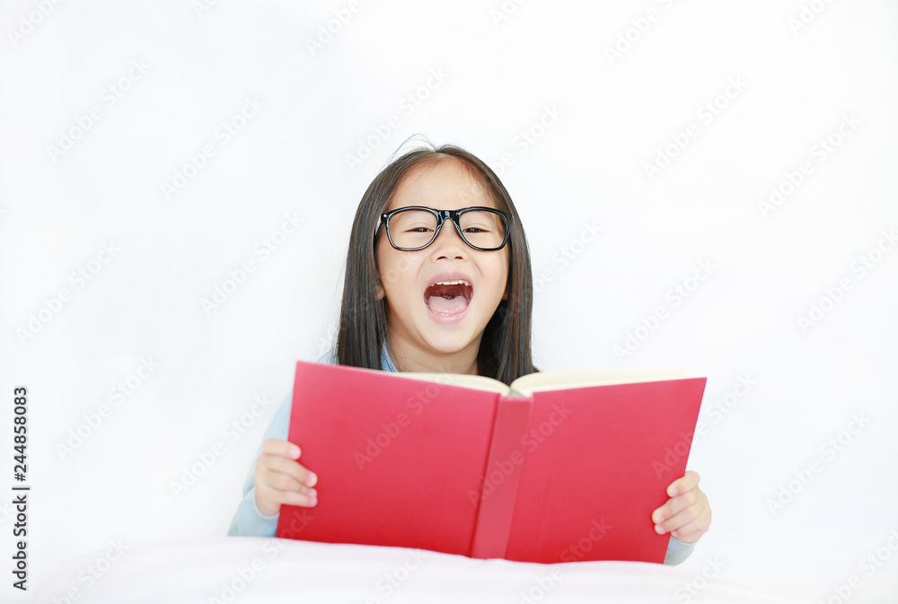 Smiling little Asian kid girl wearing glasses reading hardcover book lying on bed against white background. Smiling little Asian kid girl wearing glasses reading hardcover book lying on bed against white background.