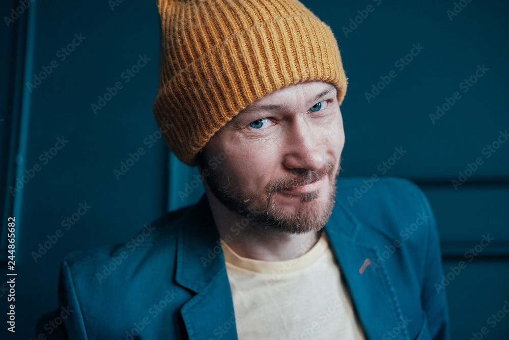 Attractive adult bearded man hipster in yellow hat looking into camera and smilingon blue wall background Attractive adult bearded man hipster in yellow hat looking into camera and smilingon blue wall background