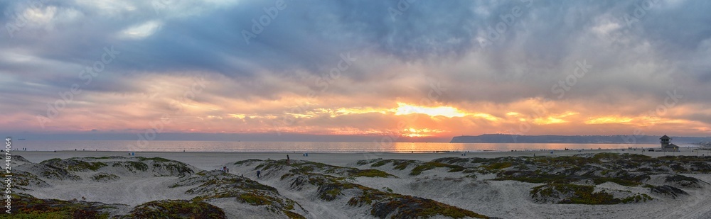Fototapeta premium Coronado Beach in San Diego by the Historic Hotel del Coronado, at sunset with unique beach sand dunes, panorama view of the Pacific Ocean, silhouettes of people walking and boats in California, Unite