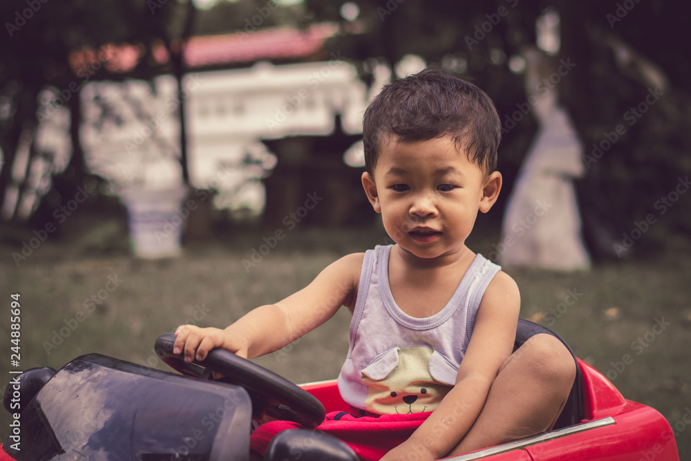 Little boy driving car with the steering wheel. Boy in a white shirt in a red toy car in the street. Little boy driving big toy car and having fun, outdoors. Little boy driving car with the steering wheel. Boy in a white shirt in a red toy car in the street. Little boy driving big toy car and having fun, outdoors.