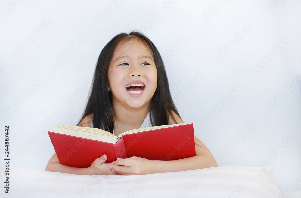 Happy little Asian kid girl reading hardcover book lying with pillow on bed against white background. Happy little Asian kid girl reading hardcover book lying with pillow on bed against white background.