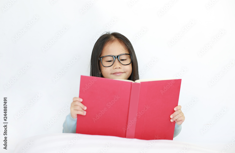 Beautiful happy little Asian kid girl reading hardcover book lying on bed against white background. Beautiful happy little Asian kid girl reading hardcover book lying on bed against white background.