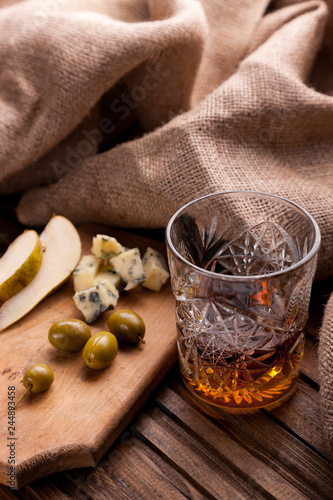 Rustic still life with whiskey and snacks. Crystal glass with a beautiful pattern on a wooden table. Snack for whiskey on crumpled paper olives  crackers  cheese  almonds  pear  apple.