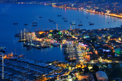 Aerial view of boats and beautiful city at night in Marmaris, Turkey. Landscape with boats in marina bay, sea, city lights, mountains, red sky, clouds at dusk. Top view from drone. Harbor at sunset
