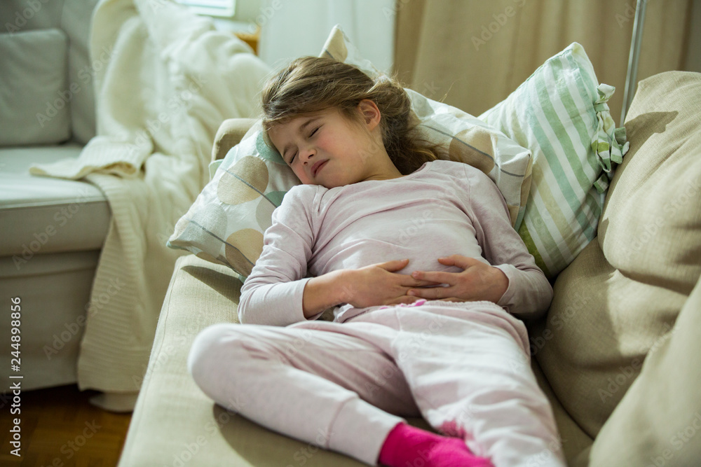Child lying on sofa in the living room with stomach pain. Hands on belly. Little girl suffering Child lying on sofa in the living room with stomach pain. Hands on belly. Little girl suffering