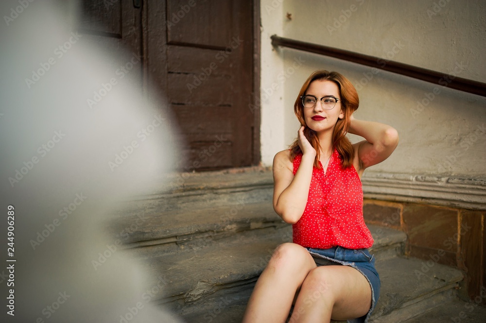 Attractive redhaired woman in eyeglasses, wear on red blouse and jeans skirt posing.