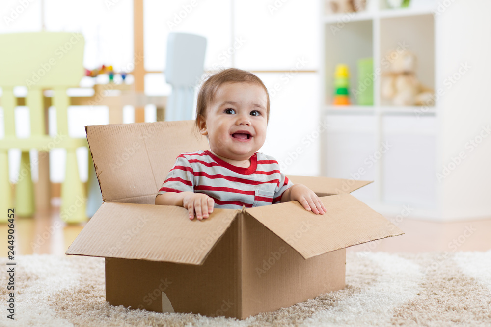 Smiling baby boy sitting inside cardboard box after moving to a new apartment - obrazy, fototapety, plakaty 