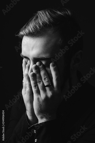 Black and white portrait of a stylish man with a beard and stylish hairdo dressed in the black shirt holding his hands on his face on the black background