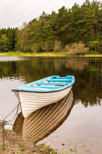 White fishing boat tied and anchored at Blessington Lake shore in Wicklow, Ireland, reflected in the calm water, on a beautiful forest background. Peaceful morning landscape,