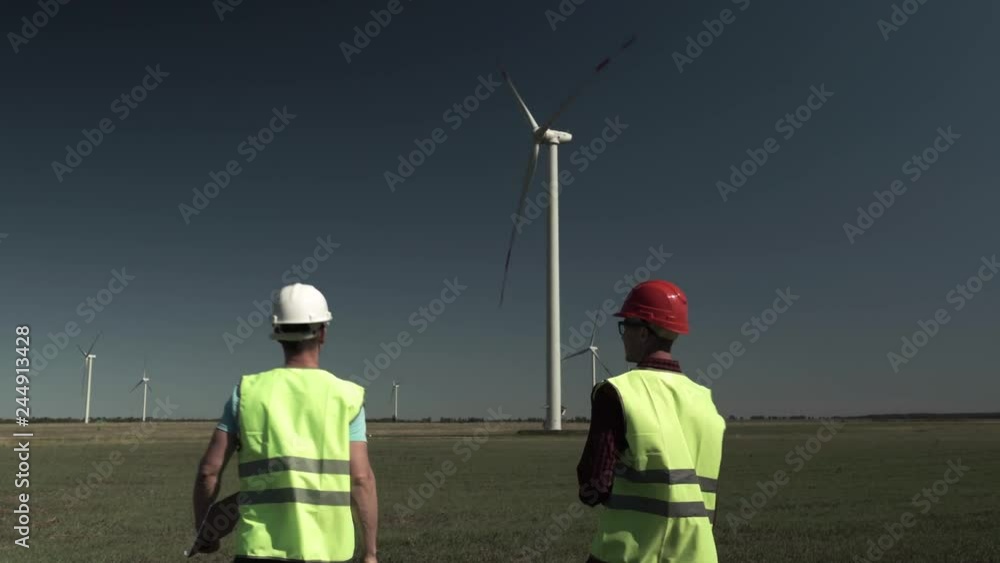 Two Young Engineers Men Approach A Windmill Power Generator And Discuss The Plan Of Its Repair In The Field On A Sunny, Clear Day Two Young Engineers Men Approach A Windmill Power Generator And Discuss The Plan Of Its Repair In The Field On A Sunny, Clear Day