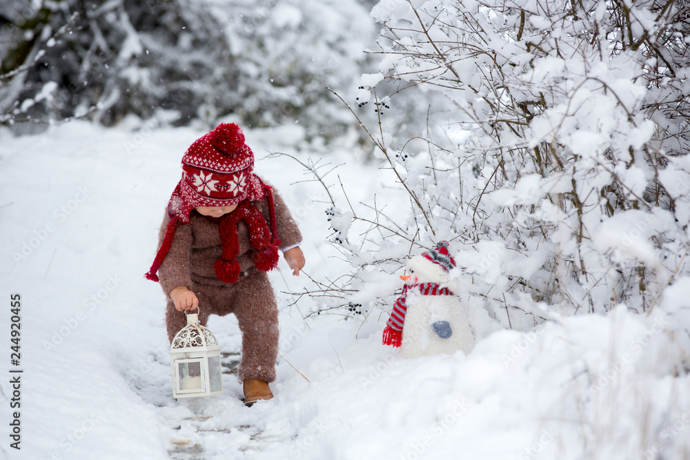 Portrait of a cute toddler baby dressed in a brown hand knitted jacket, pants, red hat and scarf, holding teddy and lantern, walks through the snowy park enjoying first snow blowing