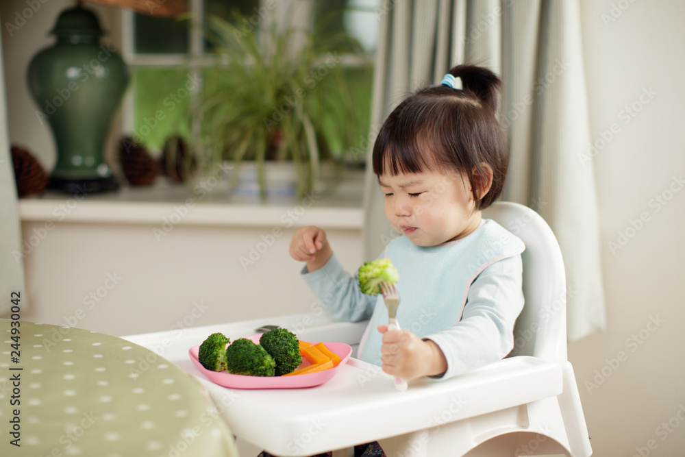 toddler girl eating healthy vegetable sitting on high chair beside a dinner table at home toddler girl eating healthy vegetable sitting on high chair beside a dinner table at home
