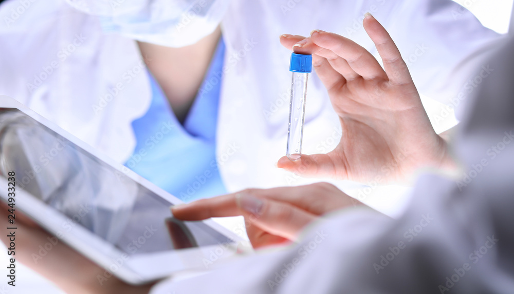 Closeup of scientific research team with clear solution in laboratory. Blonde female chemist holds test tube of glass while her colleague checks results with tablet pc. Blood test, medicine or Closeup of scientific research team with clear solution in laboratory. Blonde female chemist holds test tube of glass while her colleague checks results with tablet pc. Blood test, medicine or