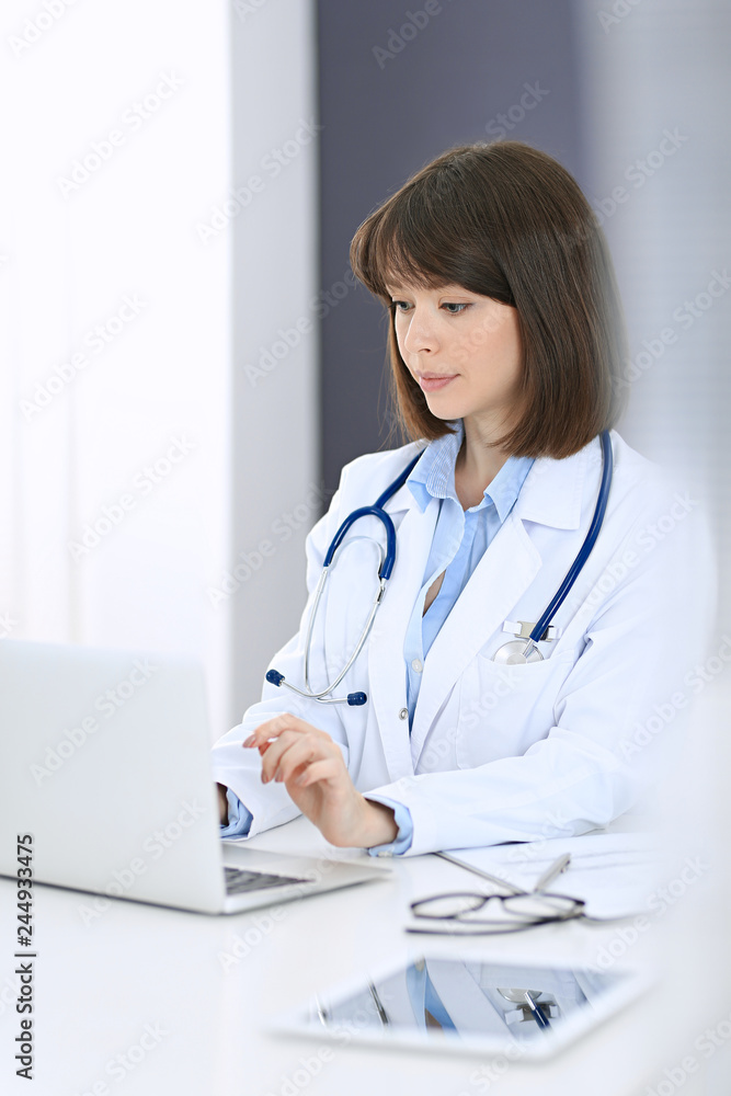 Doctor typing on laptop computer while sitting at the white table in hospital office. Physician at work. Medicine and healthcare concept Doctor typing on laptop computer while sitting at the white table in hospital office. Physician at work. Medicine and healthcare concept
